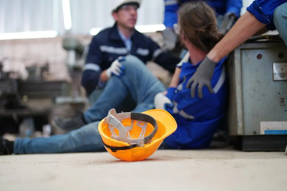 Worker injured on a factory floor, colleagues assisting. Yellow hard hat on the floor.