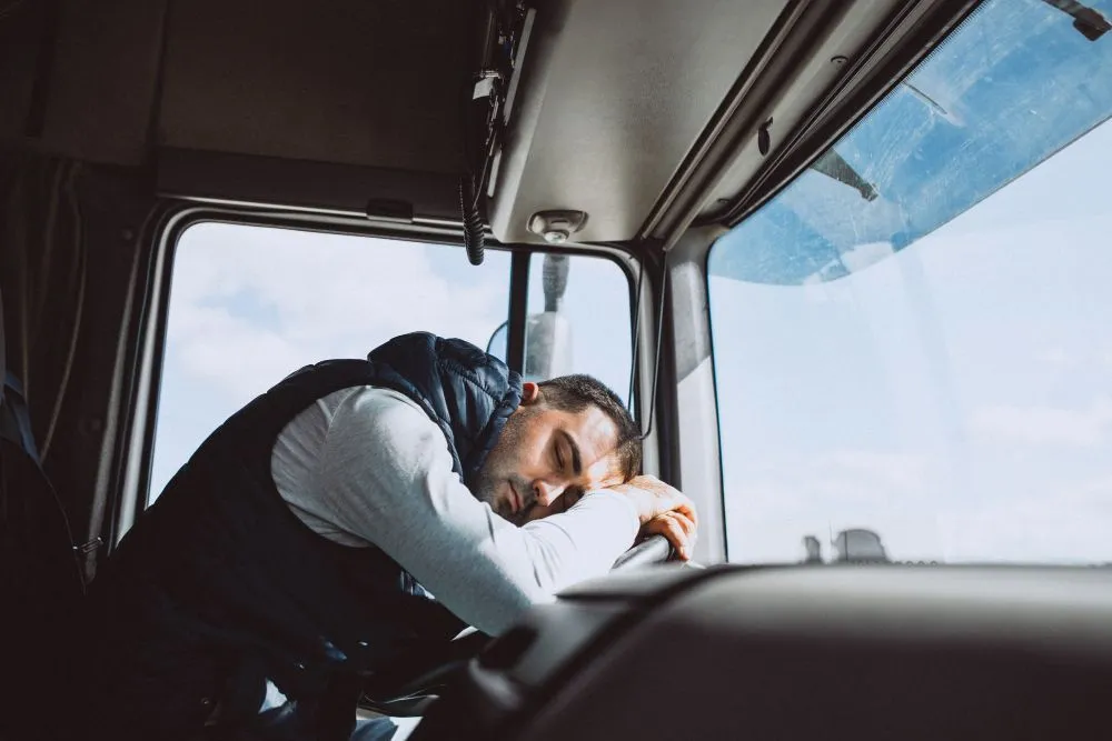 Man asleep in the driver's seat of a truck, head resting on the steering wheel, bright daylight visible outside the window.