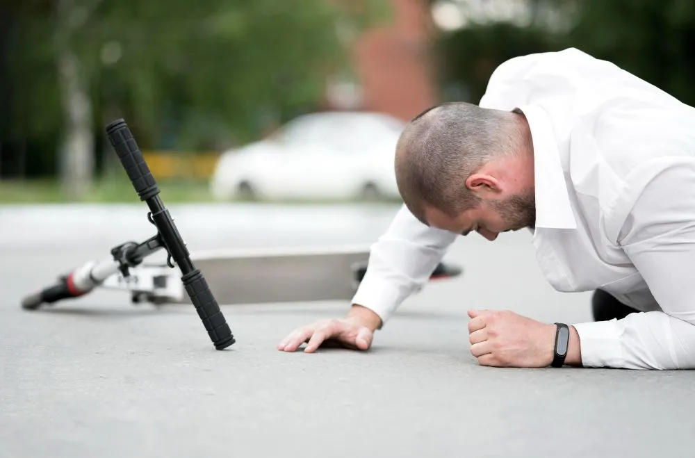 Man on ground next to fallen scooter, possibly after a fall. Asphalt setting, car in the background.