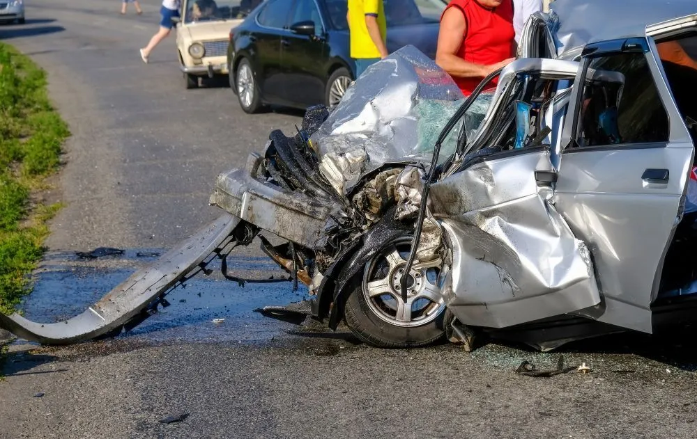 Heavily damaged silver car after a collision on a road. People stand nearby observing the wreckage.