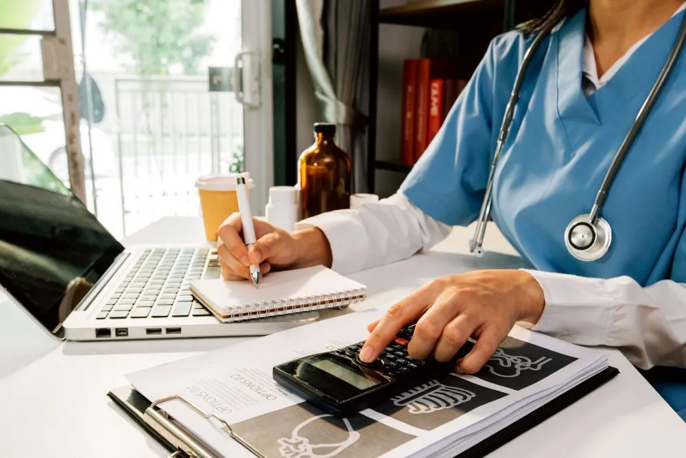 Medical professional in blue scrubs working at a desk, writing notes, using a calculator, and reviewing paperwork.