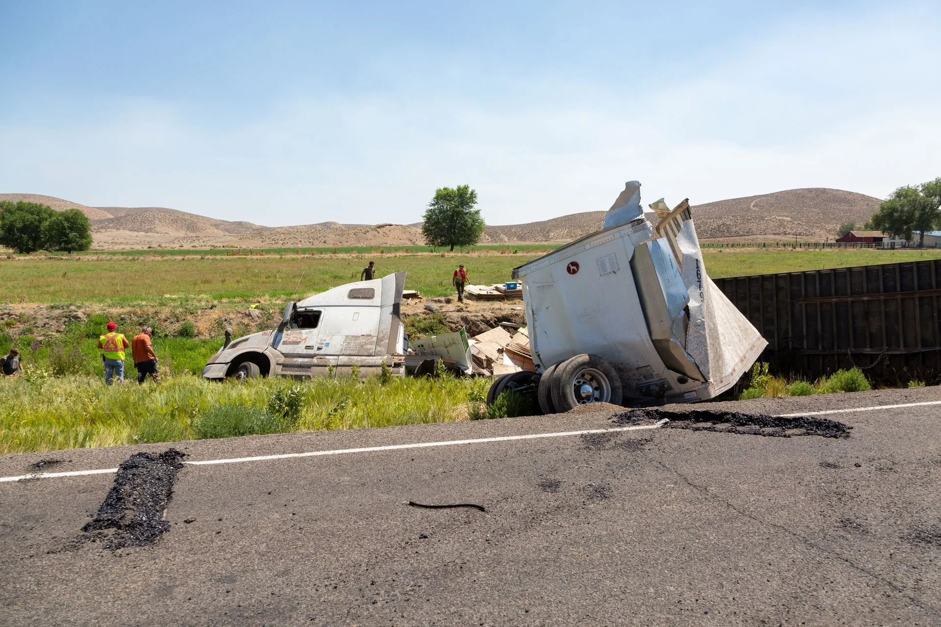 Semi-truck crashed off the road, trailer damaged. People stand nearby. Daytime.