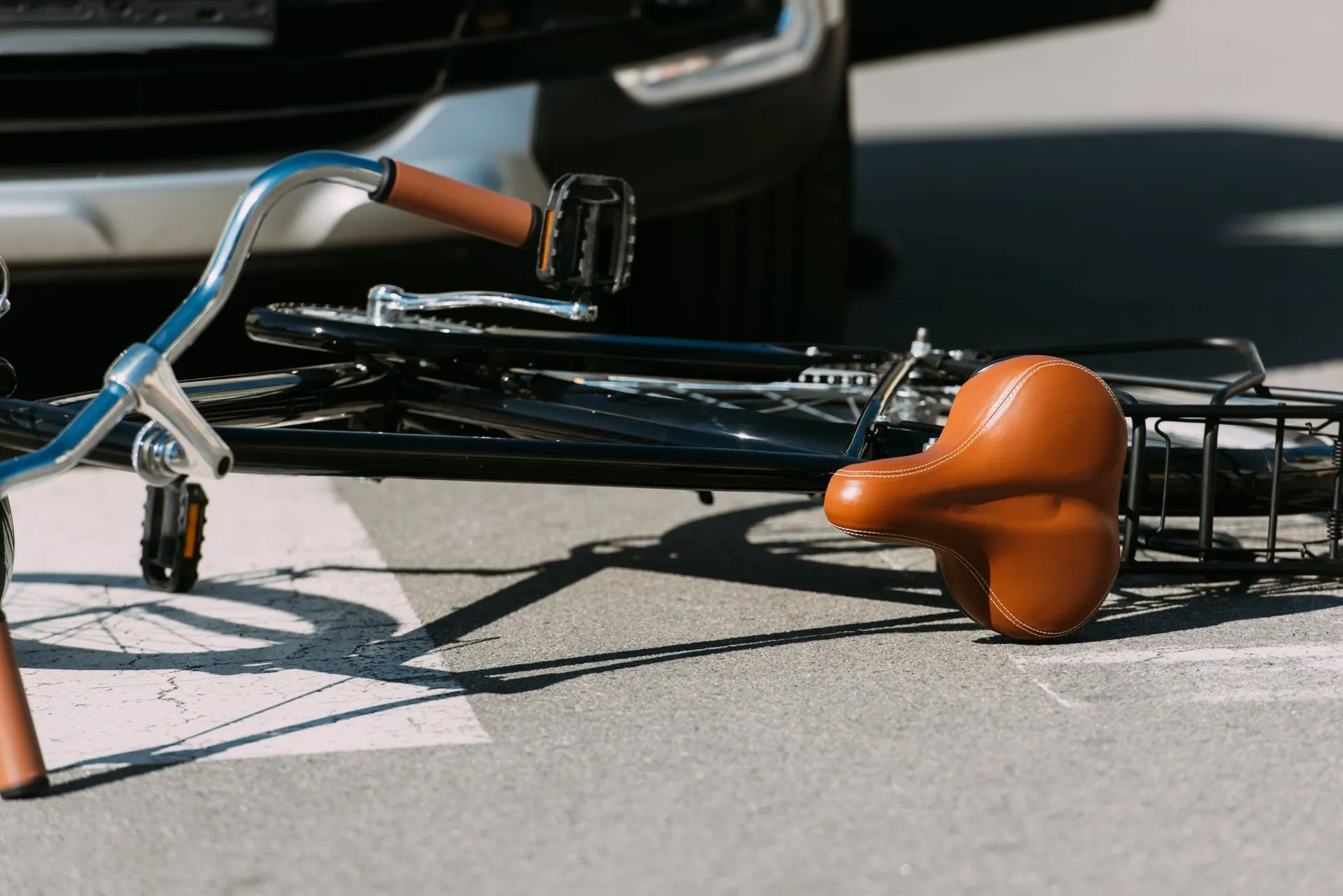 Fallen bicycle on a crosswalk next to a vehicle; brown seat, handlebars, and pedals.