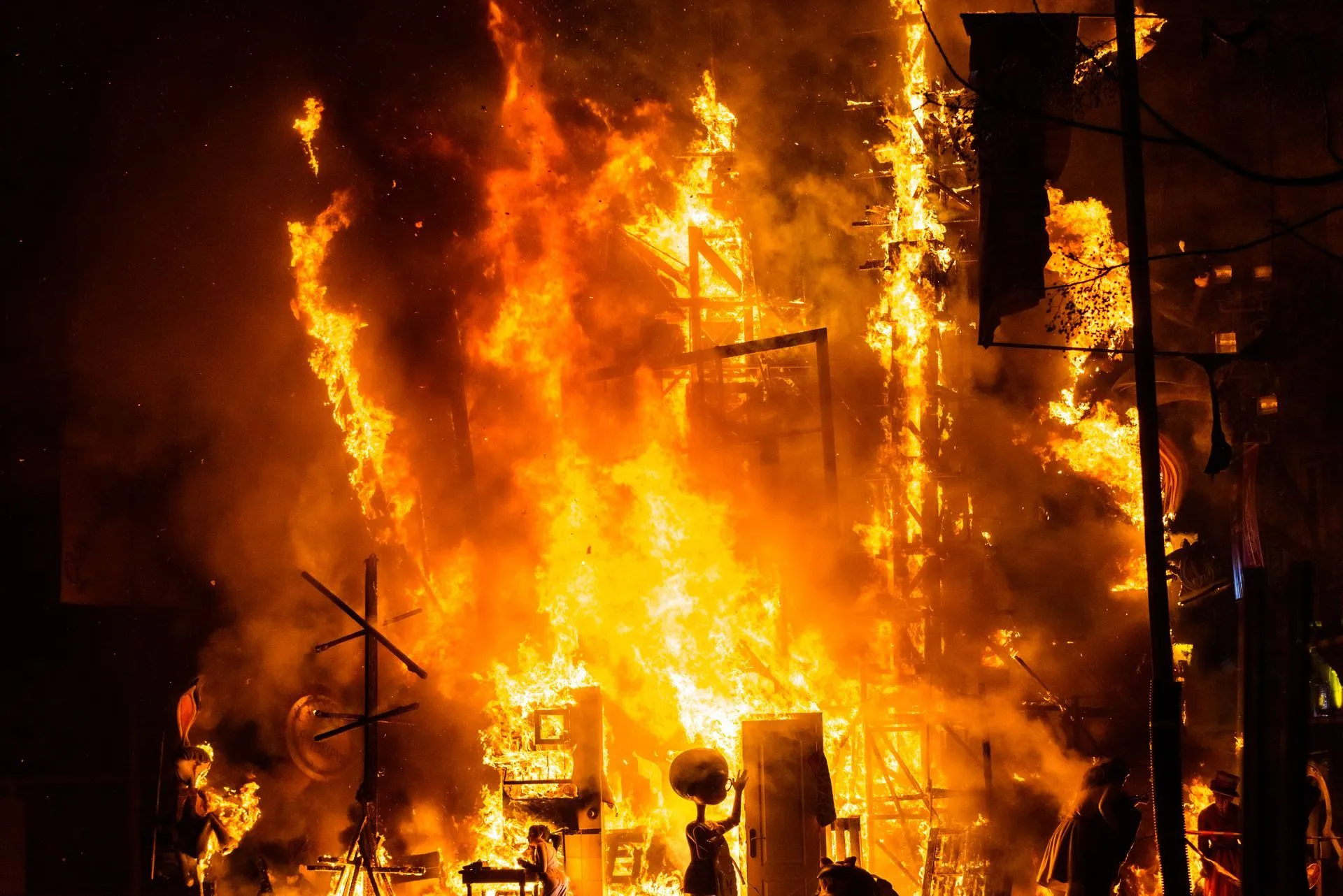 Building engulfed in flames, bright orange and yellow fire against a dark background.