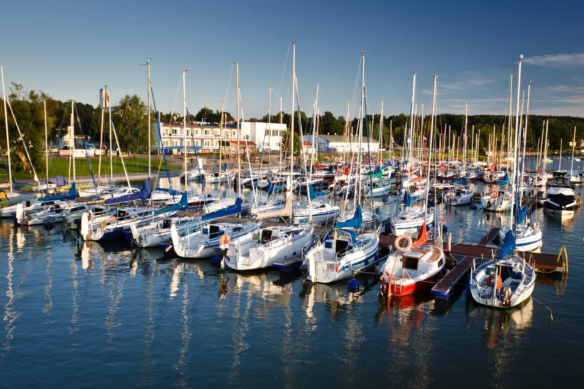 Sailboats docked at a marina on a sunny day. Blue water reflects boats and buildings.