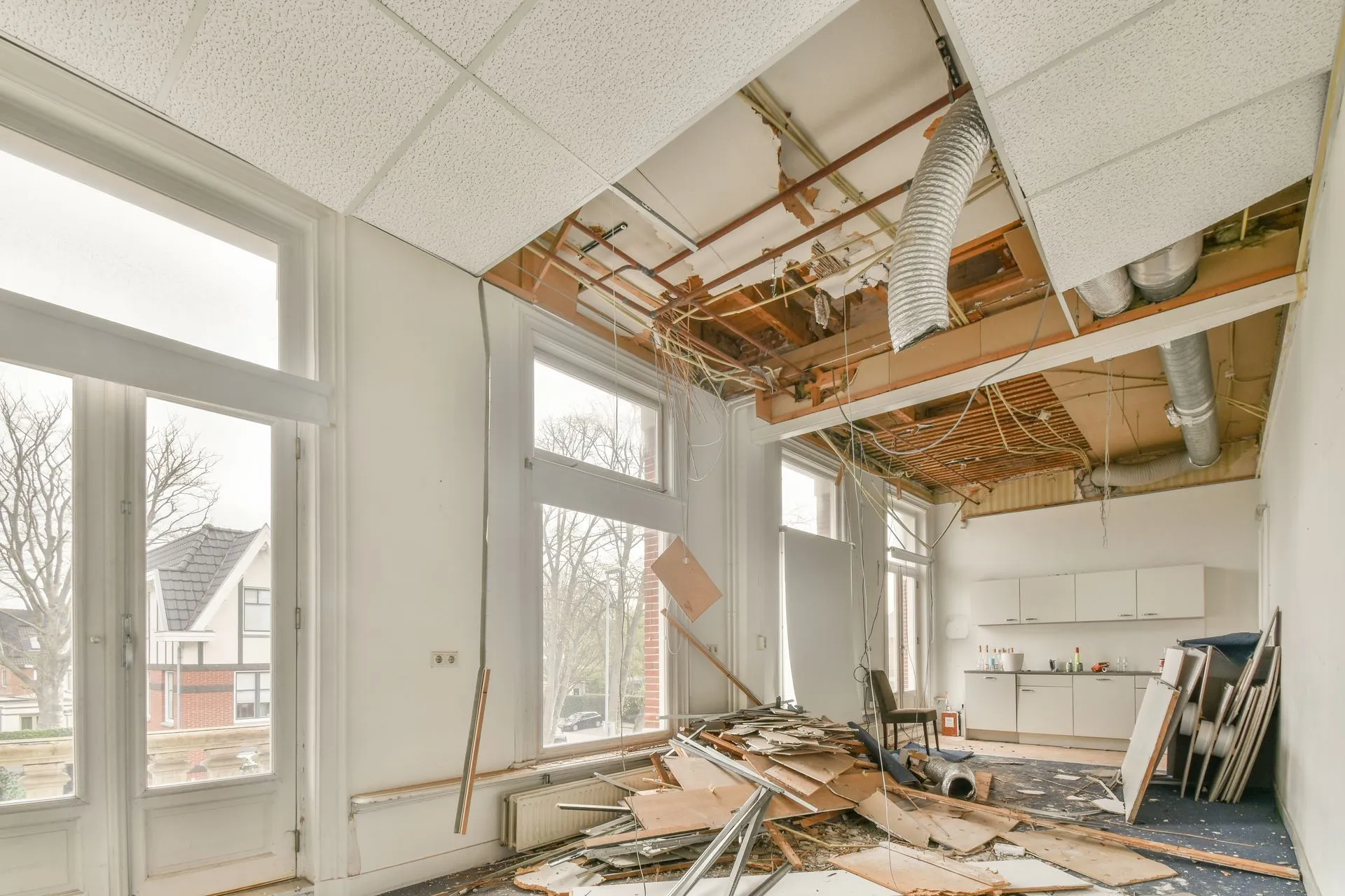 Room undergoing demolition: damaged ceiling, debris on floor, large windows.