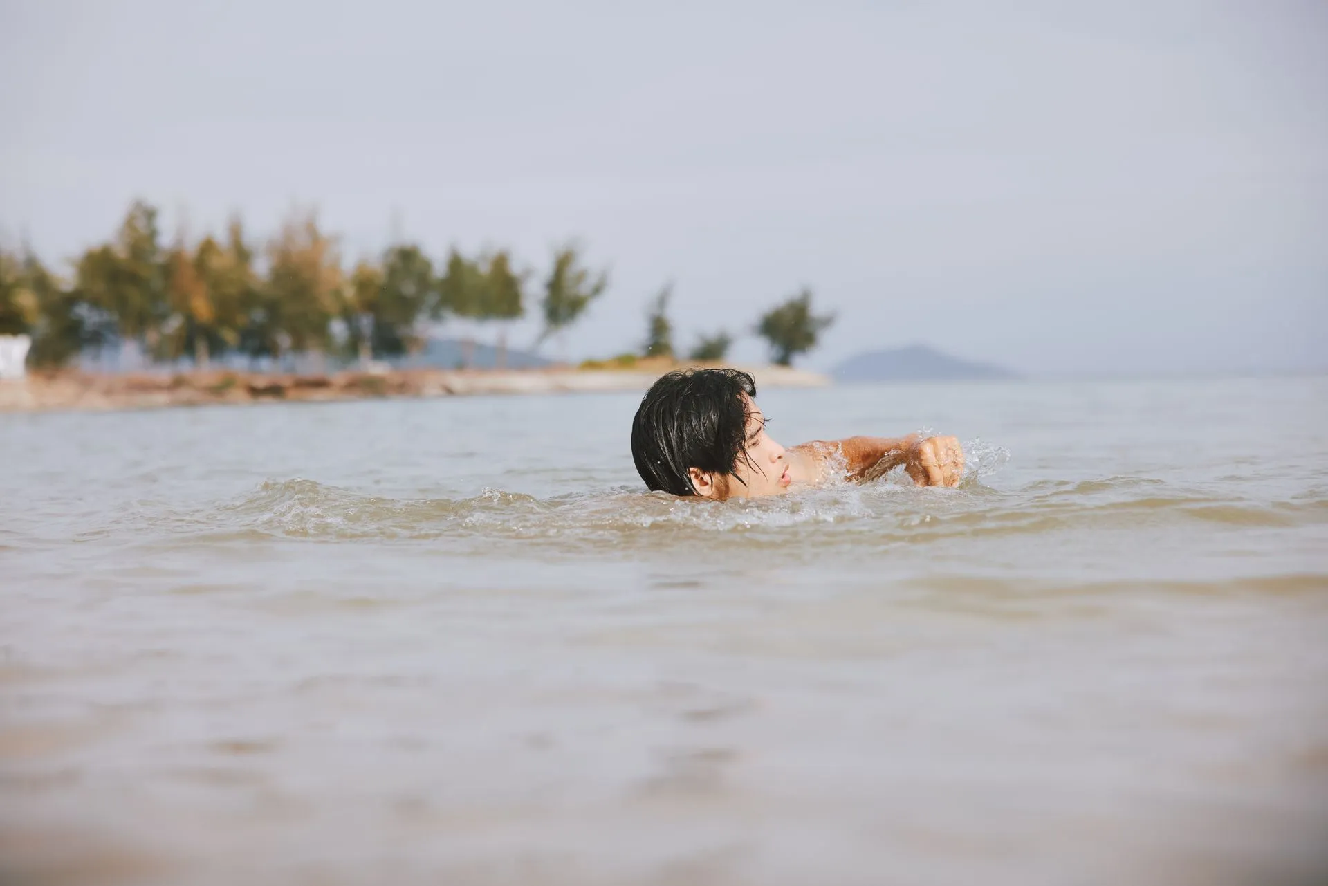 Person swimming in shallow water near a shoreline with trees, ocean, and overcast sky.