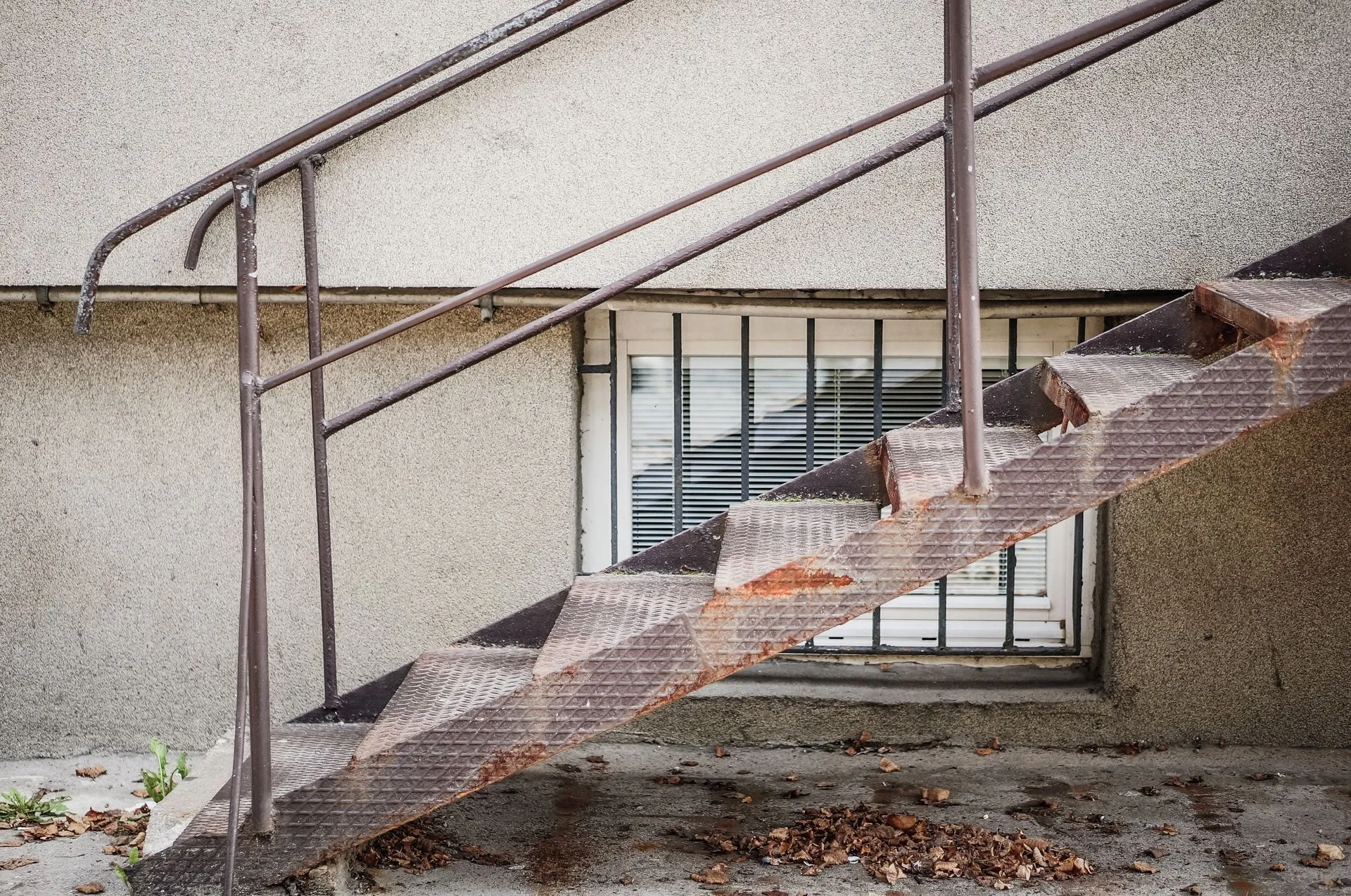 Rusty metal staircase attached to a textured beige wall, with a barred window.