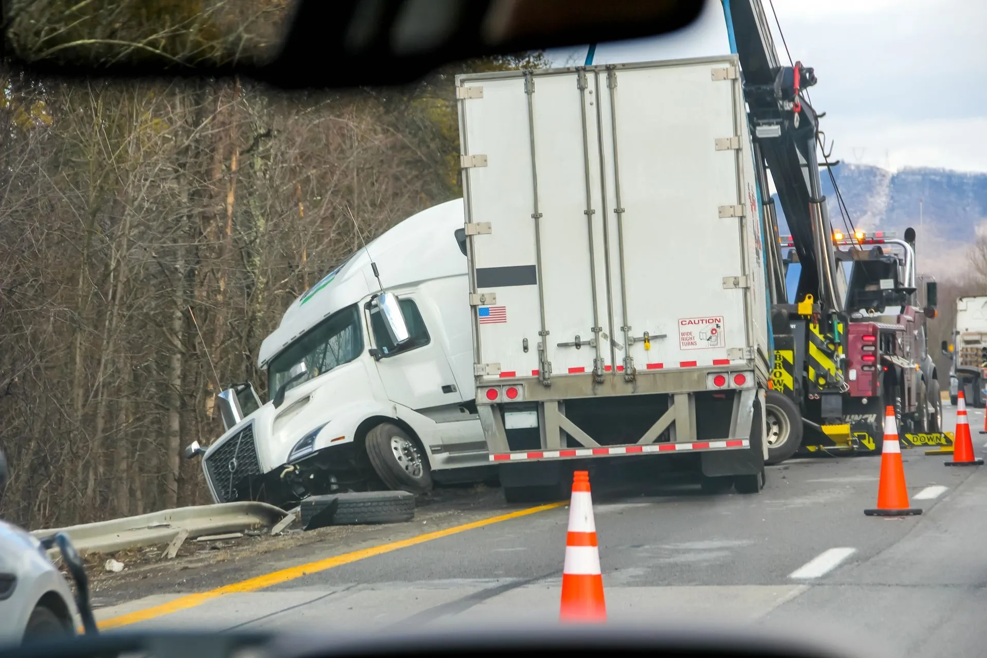 Semi-truck accident scene: white cab against guardrail, trailer in lane, tow truck present, orange cones.