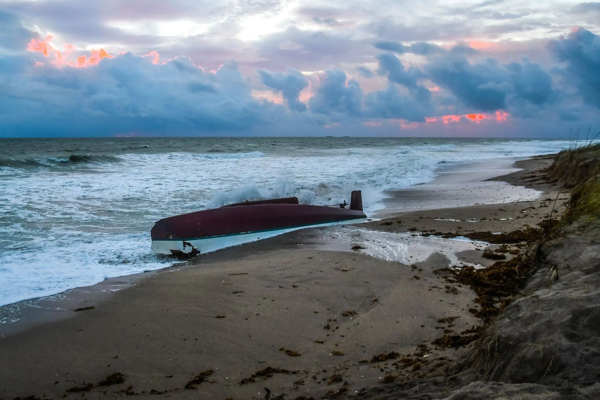 Boat washed ashore on a beach with foamy waves under a cloudy, colorful sky.